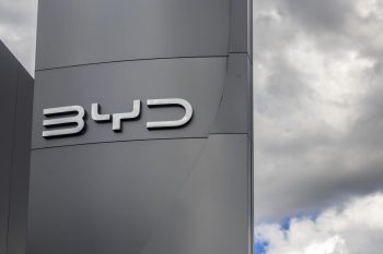 A close-up of a sleek silver BYD logo against a gray background and cloudy sky.