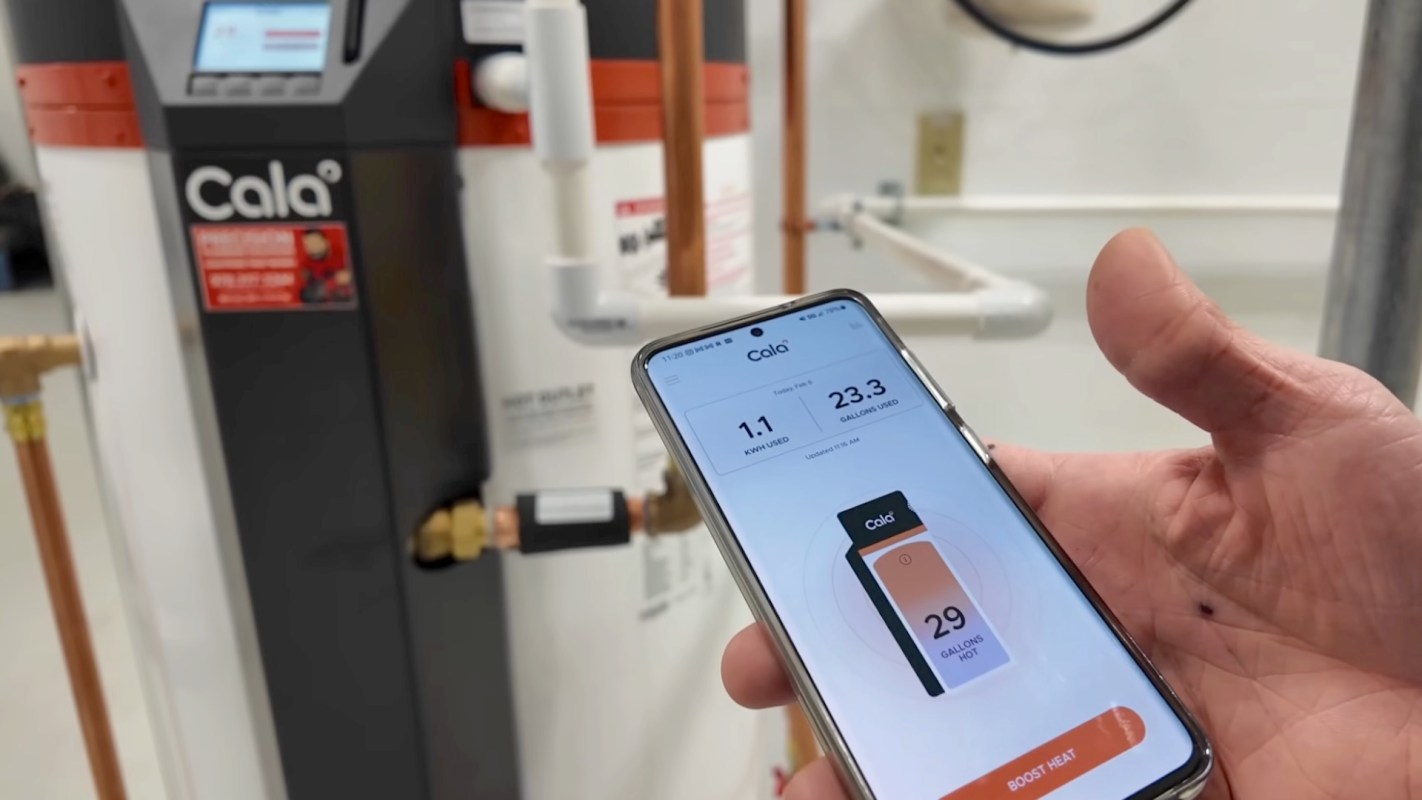 A hand holding a smartphone displays a water heater app next to a water heater unit with visible pipes.