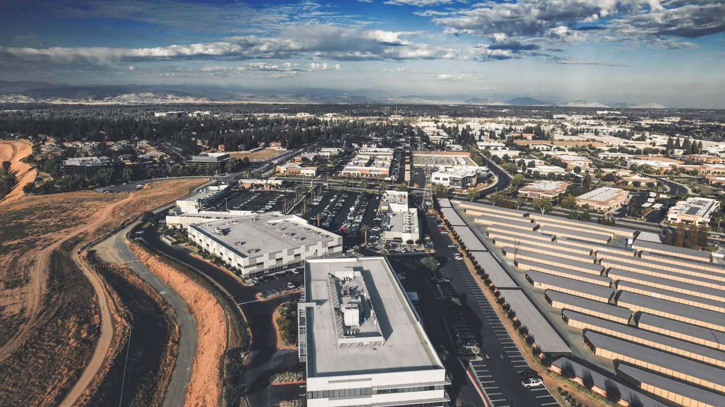 An aerial view of a suburban commercial area with buildings, parking lots, and distant mountains.