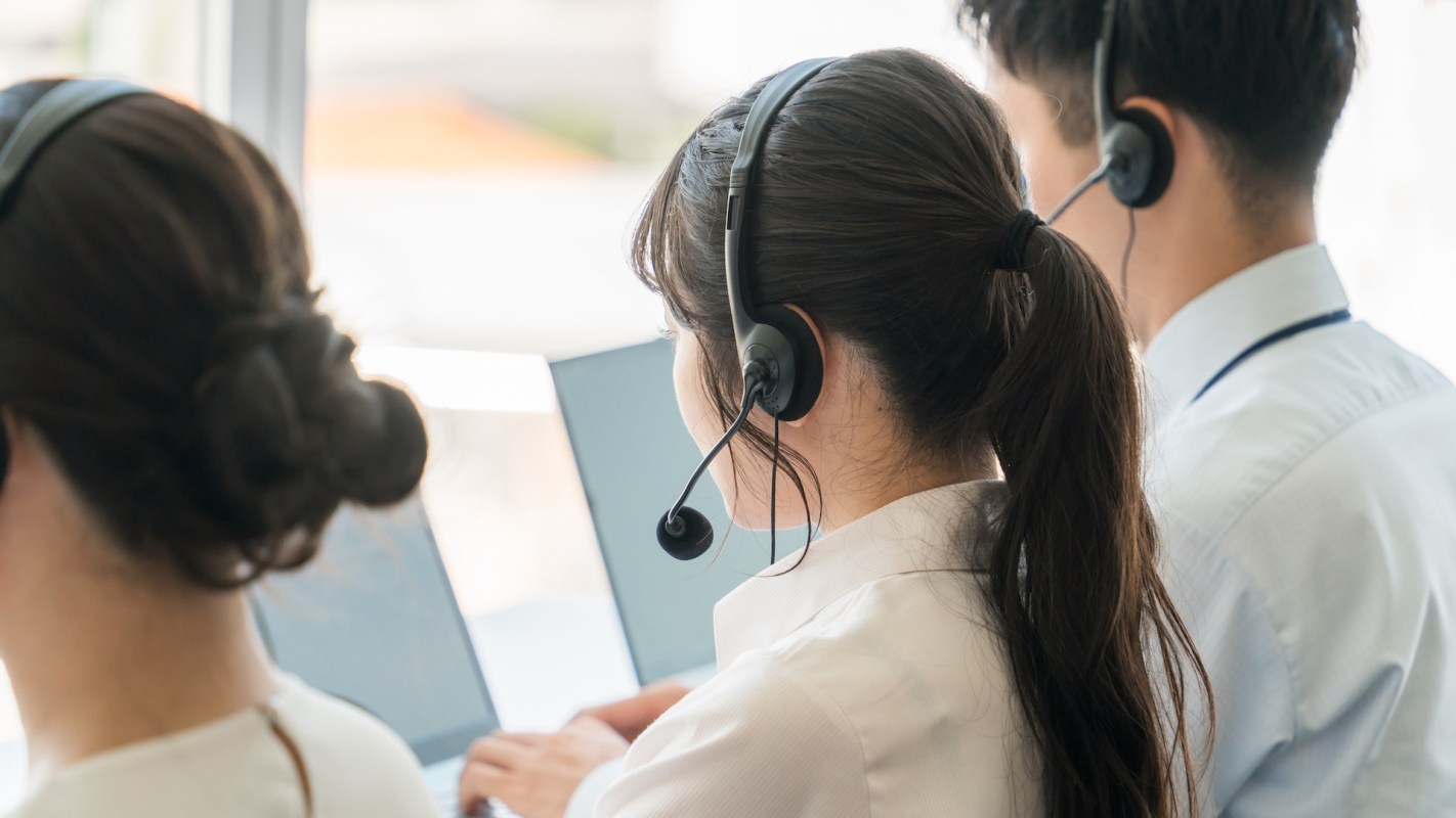 Three individuals wearing headsets work at computers in a bright office environment.