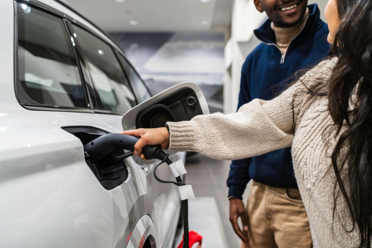 A woman plugs an electric charging cable into a white car while a man smiles in the background.