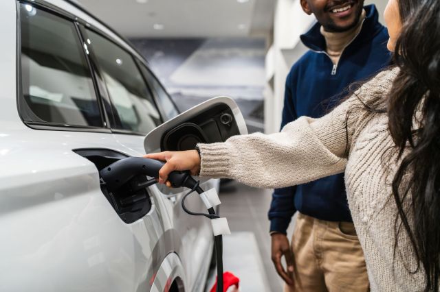 A woman plugs an electric charging cable into a white car while a man smiles in the background.