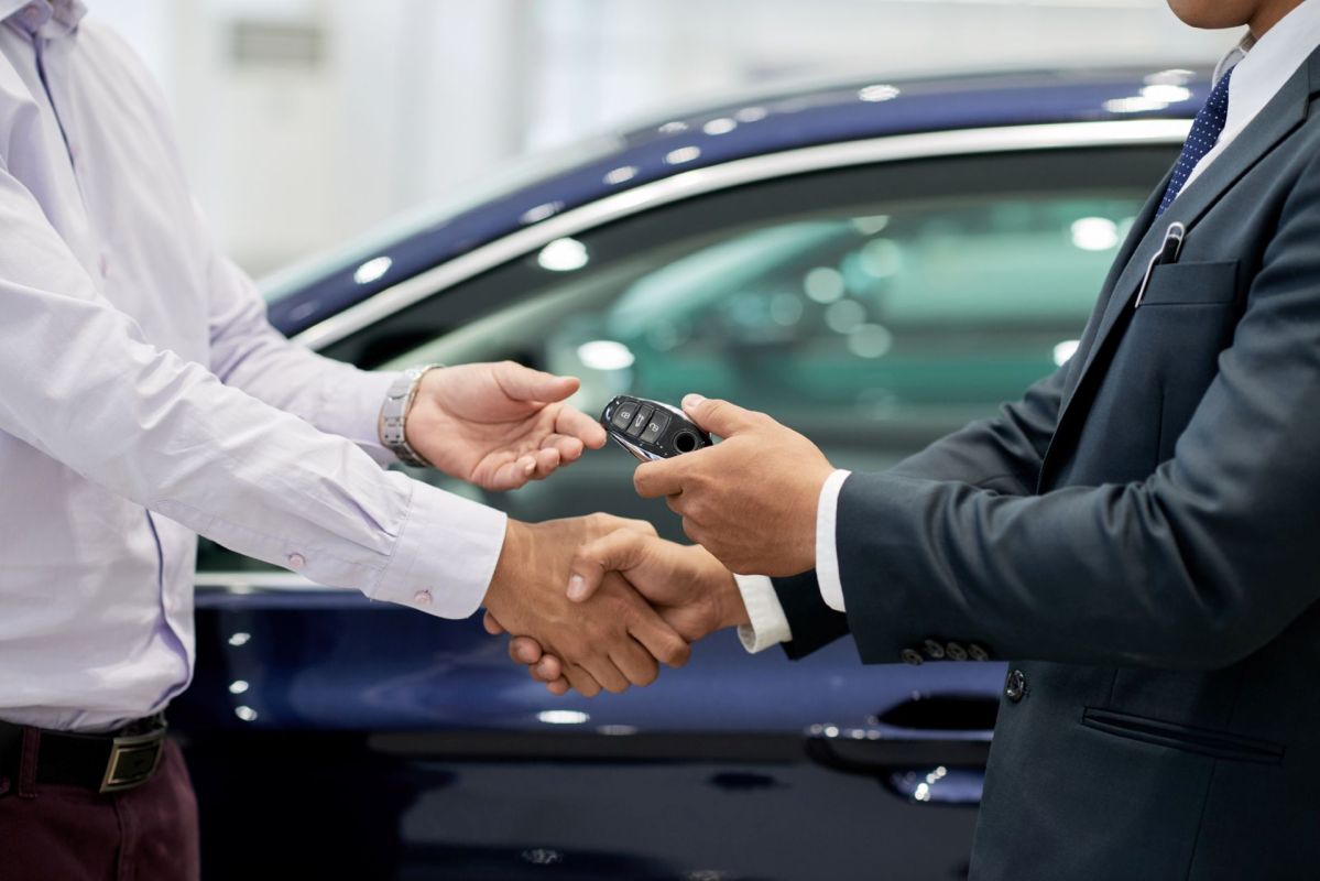 A businessman hands over car keys while shaking hands with a customer by a blue car in a dealership.