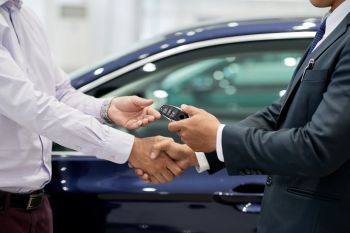 A businessman hands over car keys while shaking hands with a customer by a blue car in a dealership.