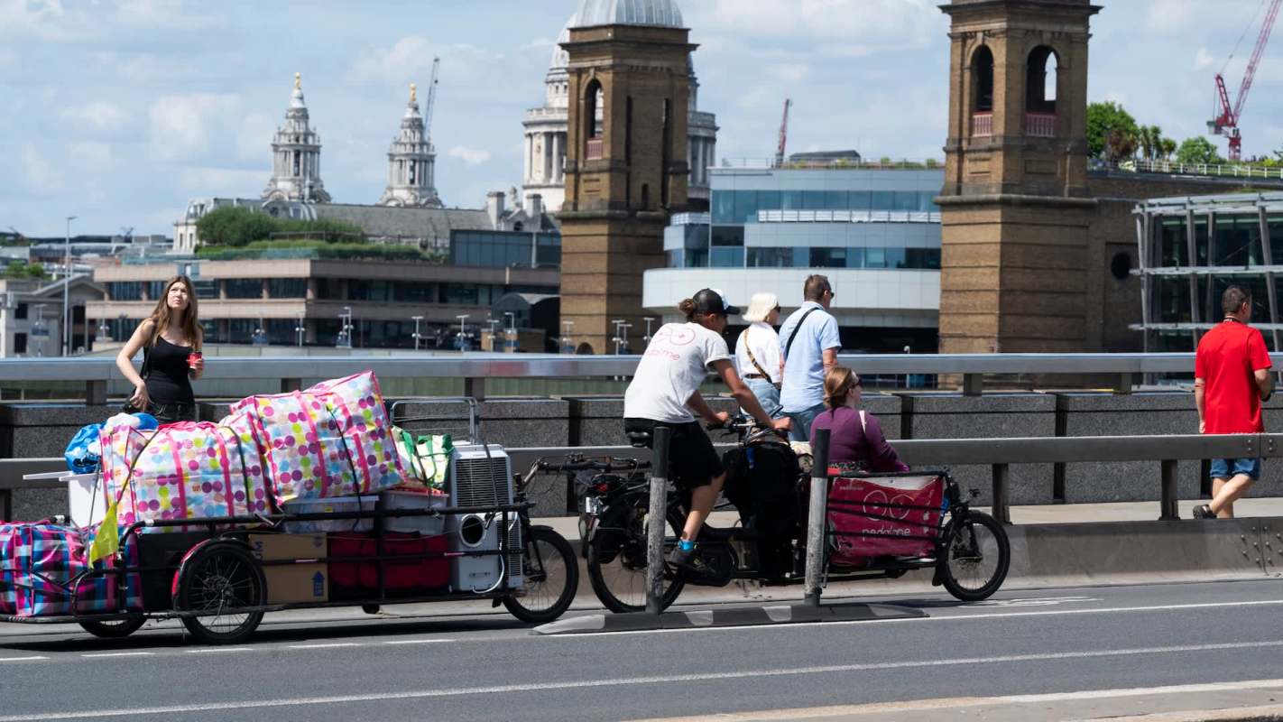 A woman stands beside colorful bags on a cart while cyclists on cargo bikes and pedestrians move along a busy urban bridge.