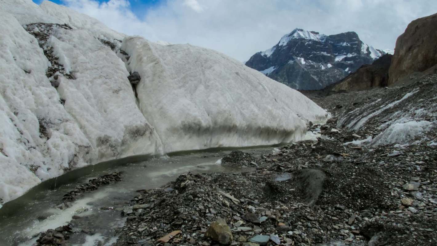 A glacier flows between rocky terrain with snow-capped mountains in the background under a cloudy sky.