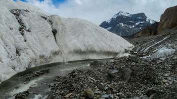 A glacier flows between rocky terrain with snow-capped mountains in the background under a cloudy sky.