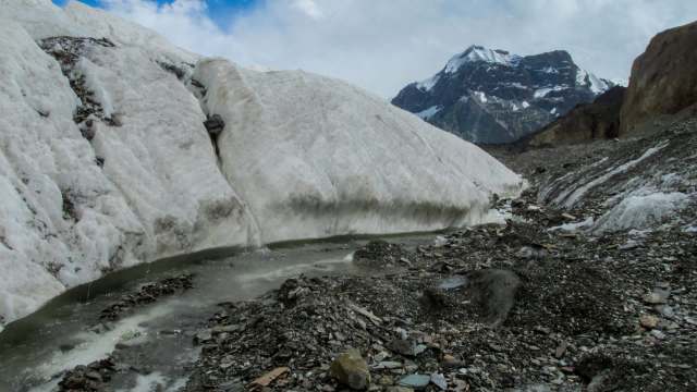 A glacier flows between rocky terrain with snow-capped mountains in the background under a cloudy sky.
