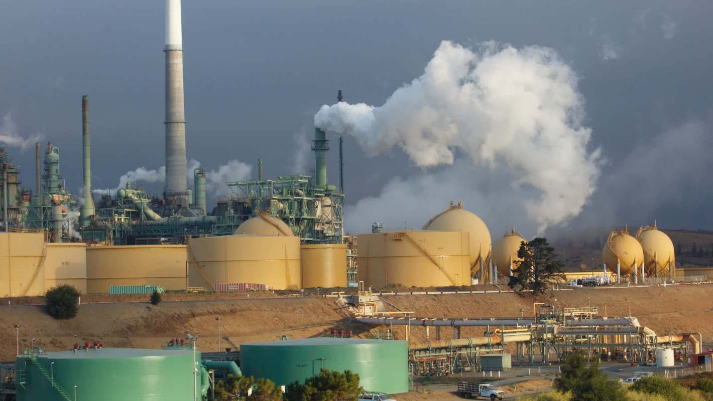 An industrial facility with large storage tanks and smokestacks emitting steam and smoke against a cloudy sky.