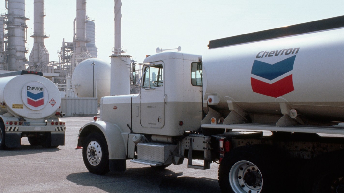 Two Chevron tanker trucks are parked in an industrial facility with large storage tanks and smokestacks in the background.
