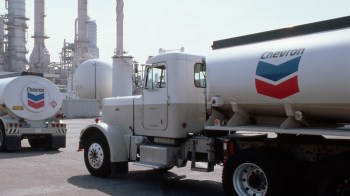 Two Chevron tanker trucks are parked in an industrial facility with large storage tanks and smokestacks in the background.