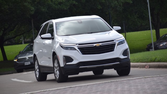 A white Chevrolet SUV drives on a paved road with green grass and trees in the background.