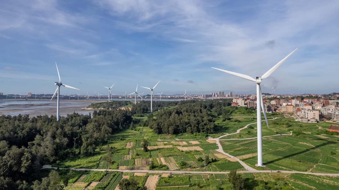 A panoramic view of wind turbines adjacent to green fields and urban buildings under a clear blue sky.