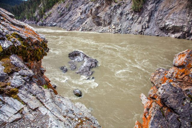 Rocky riverbank with moss and lichen, flowing brown river, surrounded by steep rocky cliffs and green forest.