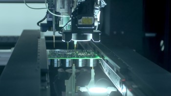 A robotic arm assembles electronic components on a green circuit board in a dark workspace.