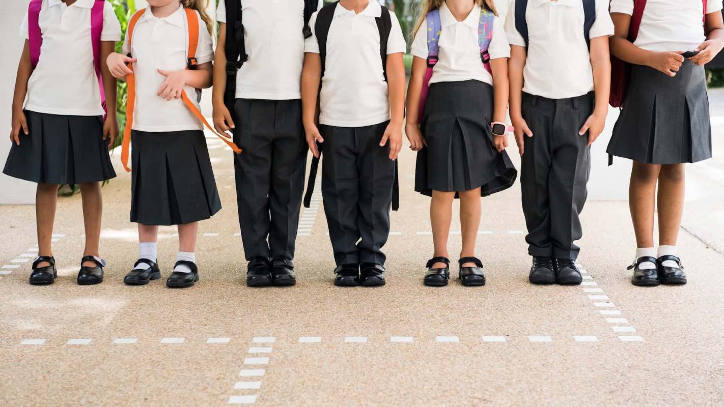 A line of schoolchildren in uniforms and backpacks standing on a playground with a textured ground.