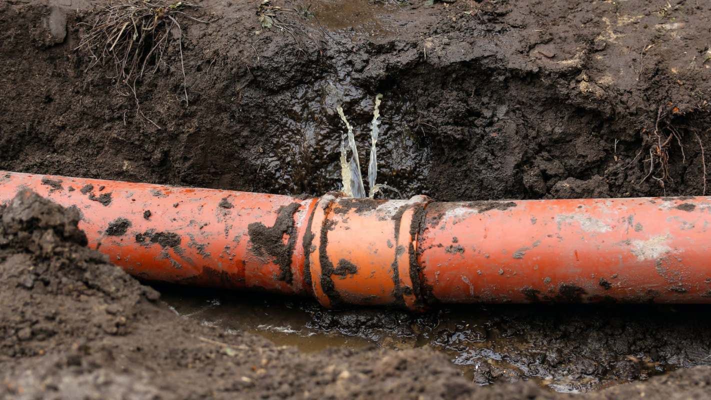 A damaged orange pipe leaks water into a muddy trench.