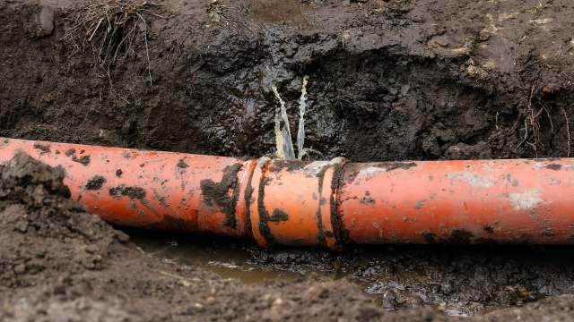 A damaged orange pipe leaks water into a muddy trench.