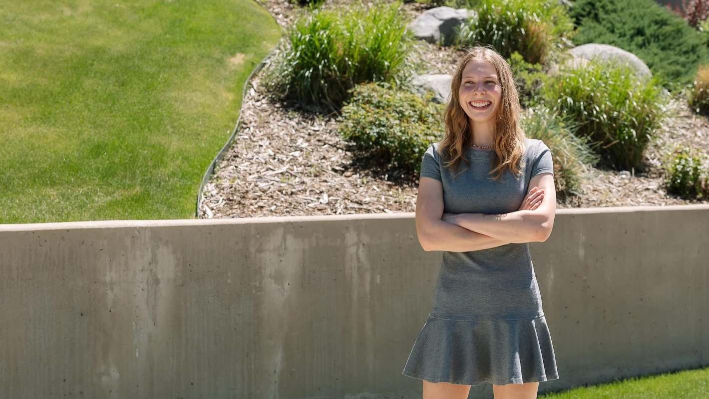 A smiling young woman stands with arms crossed in a grassy outdoor setting.