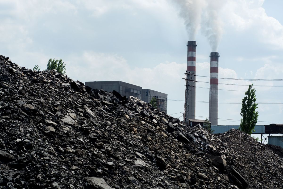 A coal mound in the foreground with industrial smokestacks emitting white smoke in the background under a cloudy sky.