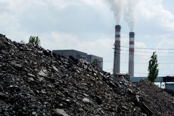 A coal mound in the foreground with industrial smokestacks emitting white smoke in the background under a cloudy sky.