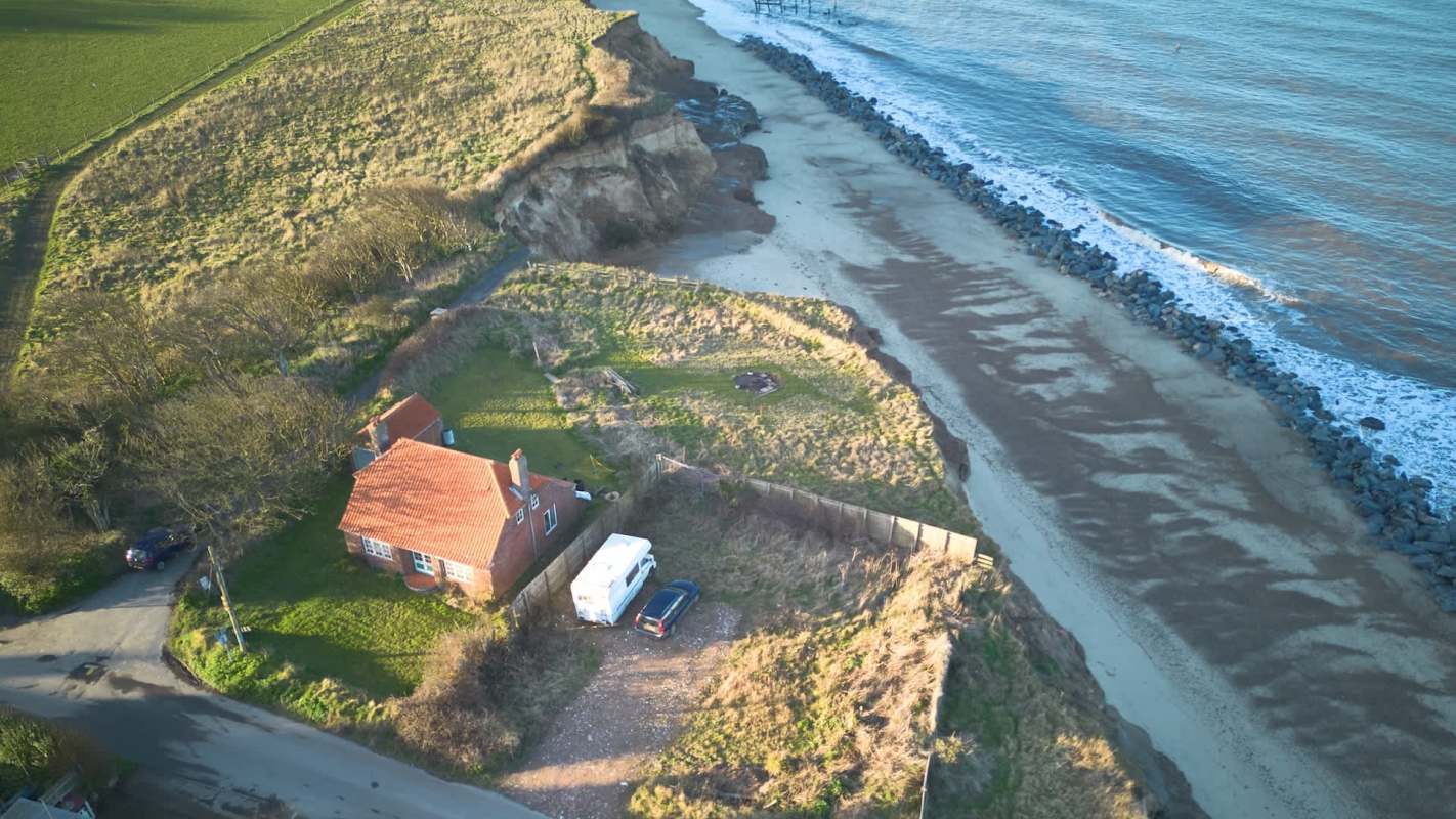 Aerial view of a coastal area featuring a house, a vehicle, and a sandy beach along the shore.