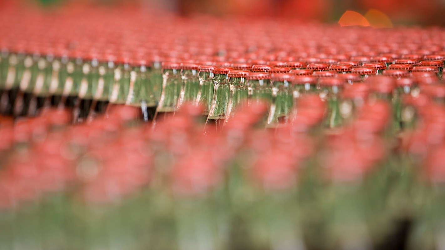 A close-up view of numerous green glass bottles with red caps arranged in a blurred pattern.