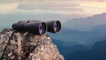 A pair of binoculars rests on a rocky surface overlooking a misty mountain landscape at sunset.