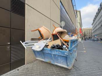 A blue dumpster overflowing with discarded furniture and debris next to a modern building.