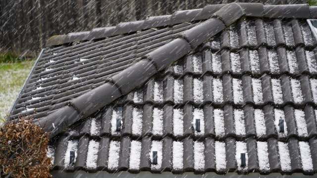 A close-up of a sloped roof covered with hail as snow falls in the background.