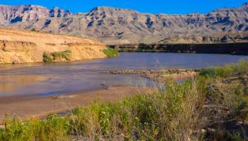 A low-flowing river meanders through rocky terrain with layered hills under a clear blue sky.