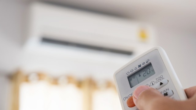 A person holds an air conditioner remote, pointing it toward a wall-mounted unit in a softly lit room.