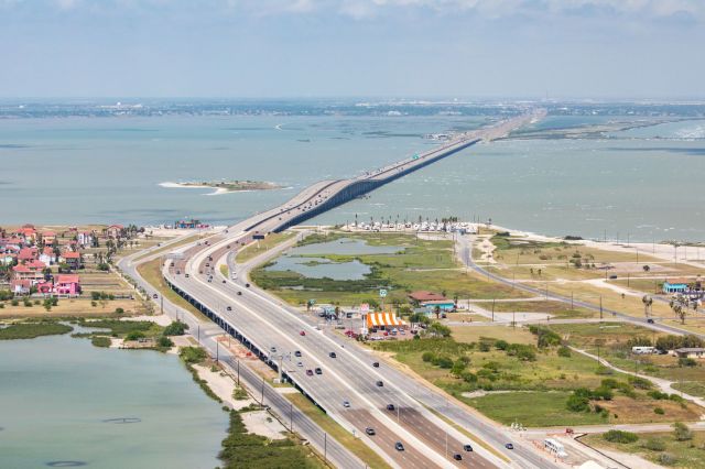 Aerial view of a highway bridge connecting two coastal areas, with water and green landscapes nearby.