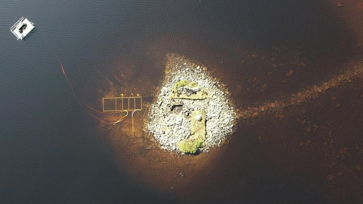An aerial view of a small rocky island in dark water, with a structure and a pathway leading to it.