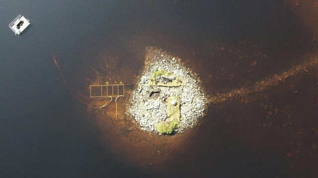 An aerial view of a small rocky island in dark water, with a structure and a pathway leading to it.