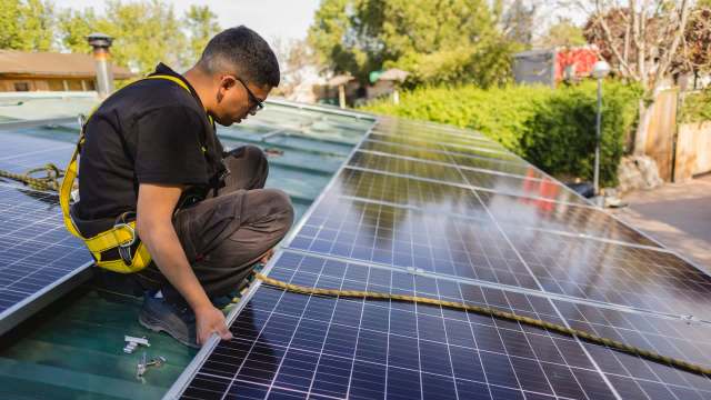A technician installs solar panels on a rooftop, adjusting the panels while secured with a safety harness.