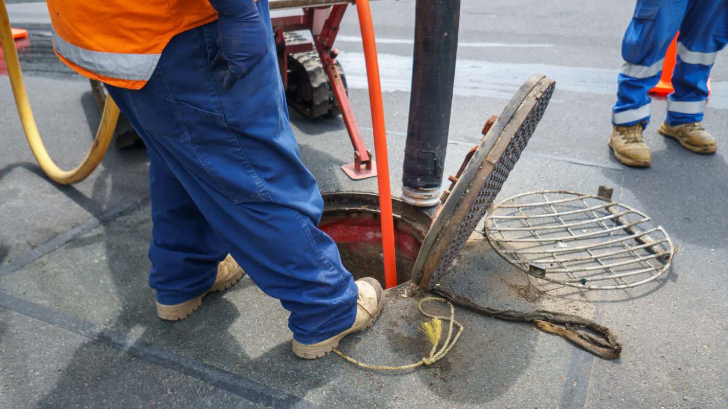 A worker in an orange vest uses a hose in a manhole while another worker stands nearby on a city street.