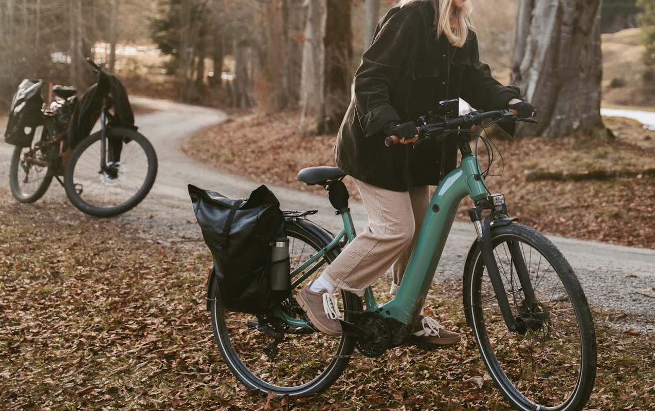 A woman in a black jacket rides a teal electric bike along a leaf-covered path with another bike in the background.