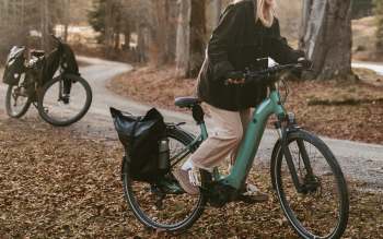 A woman in a black jacket rides a teal electric bike along a leaf-covered path with another bike in the background.