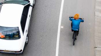 A cyclist in a blue shirt and orange helmet rides alongside a white car on a gray road.