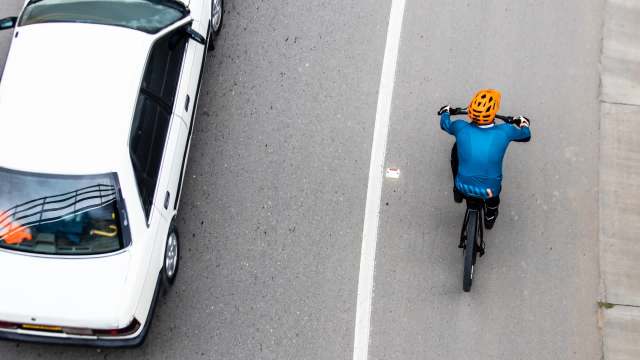 A cyclist in a blue shirt and orange helmet rides alongside a white car on a gray road.