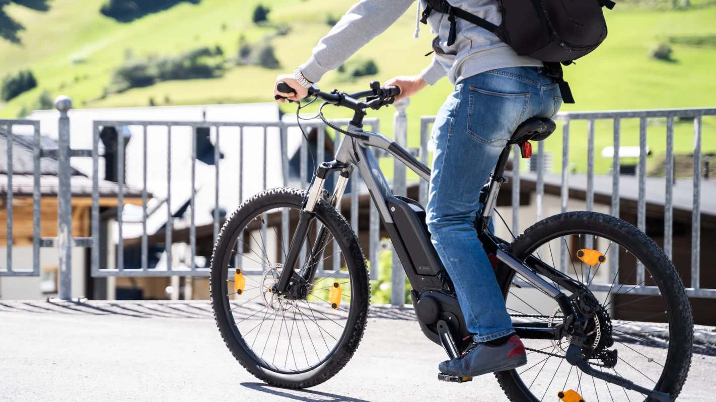 A person in a gray hoodie riding an electric mountain bike near a gate with green hills in the background.