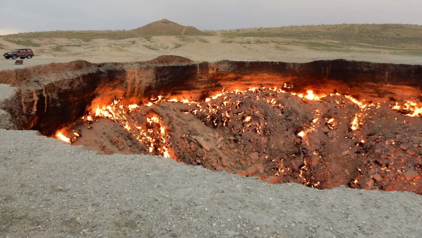 A large fiery crater in a barren landscape, the famous "Door to Hell" in Turkmenistan, with a distant vehicle parked nearby.
