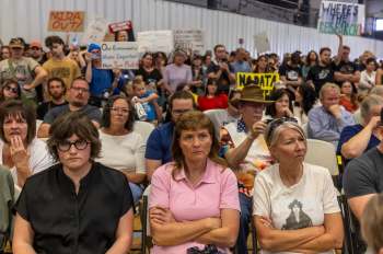 A diverse crowd of people seated, showing concern and holding protest signs about data centers at a community meeting.