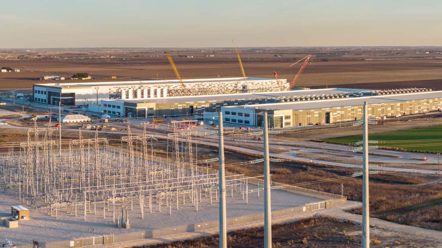 A large industrial facility under construction with power lines and electrical infrastructure in the foreground.