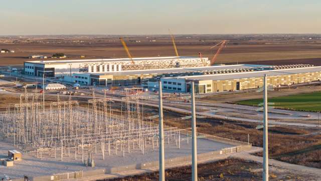 A large industrial facility under construction with power lines and electrical infrastructure in the foreground.