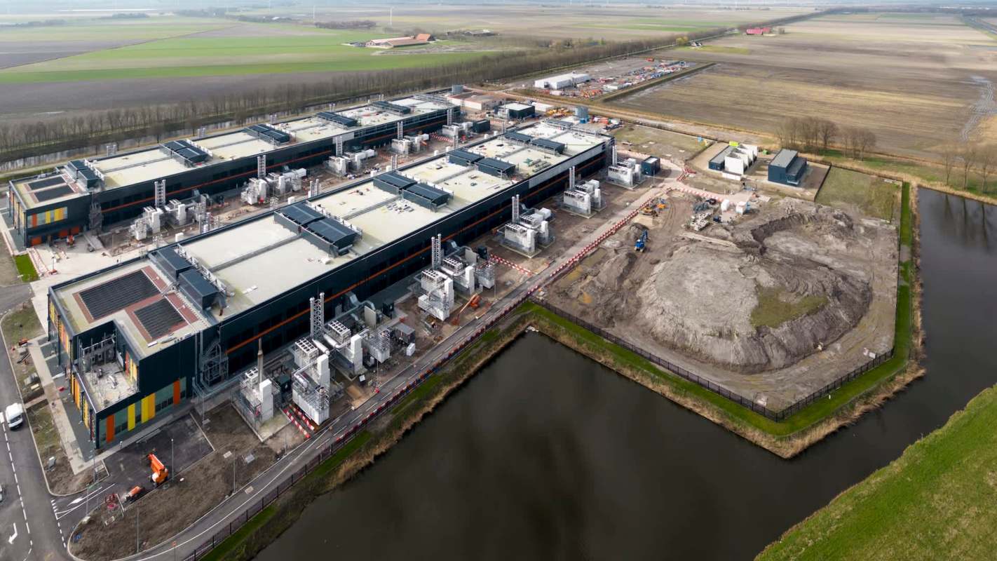 An aerial view of a large data center with construction sites and green fields surrounding it.