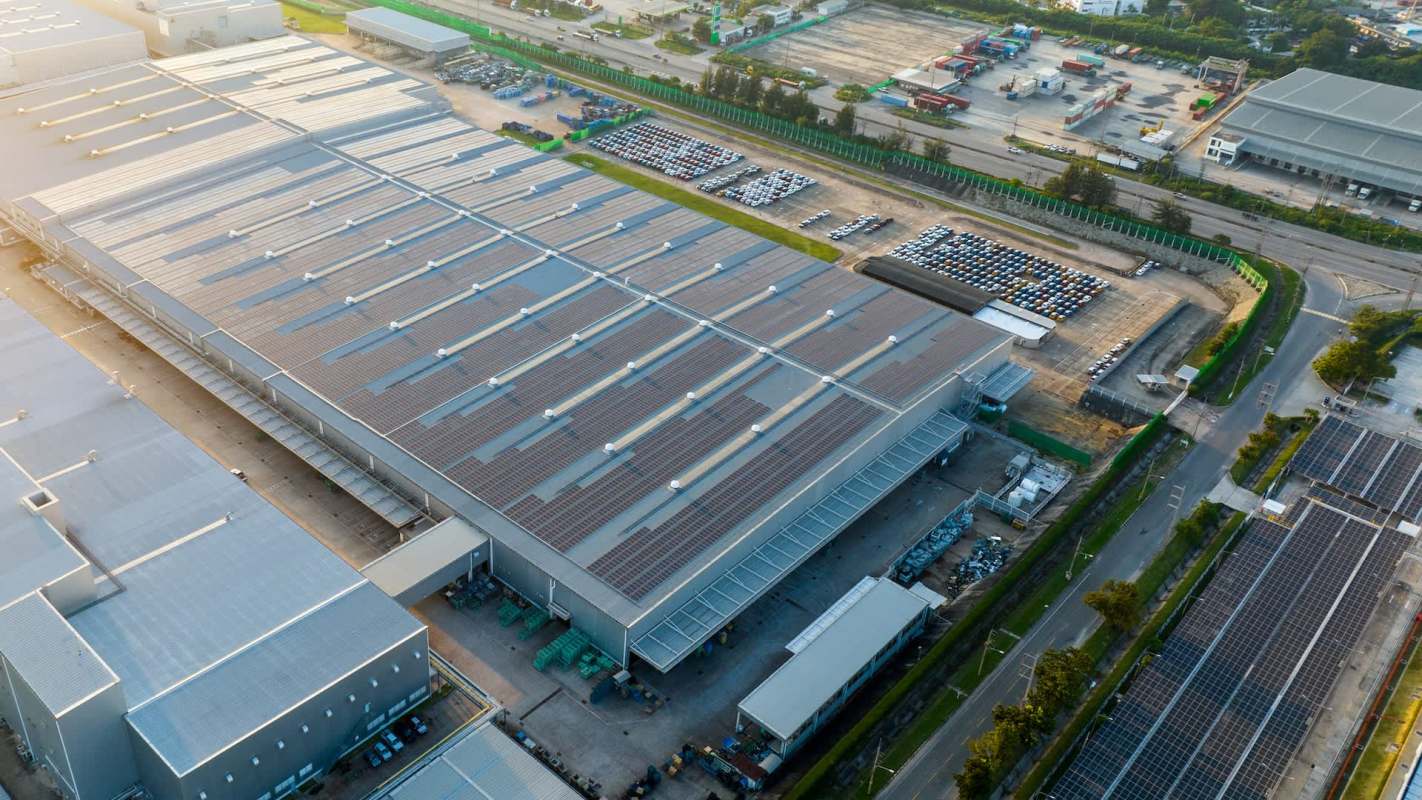 An aerial view of a large industrial building with solar panels, surrounded by parked cars and open land.