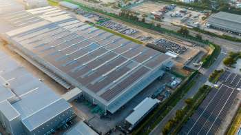 An aerial view of a large industrial building with solar panels, surrounded by parked cars and open land.