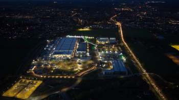 An aerial view of a brightly lit industrial area at night, with traffic flowing along a winding road.
